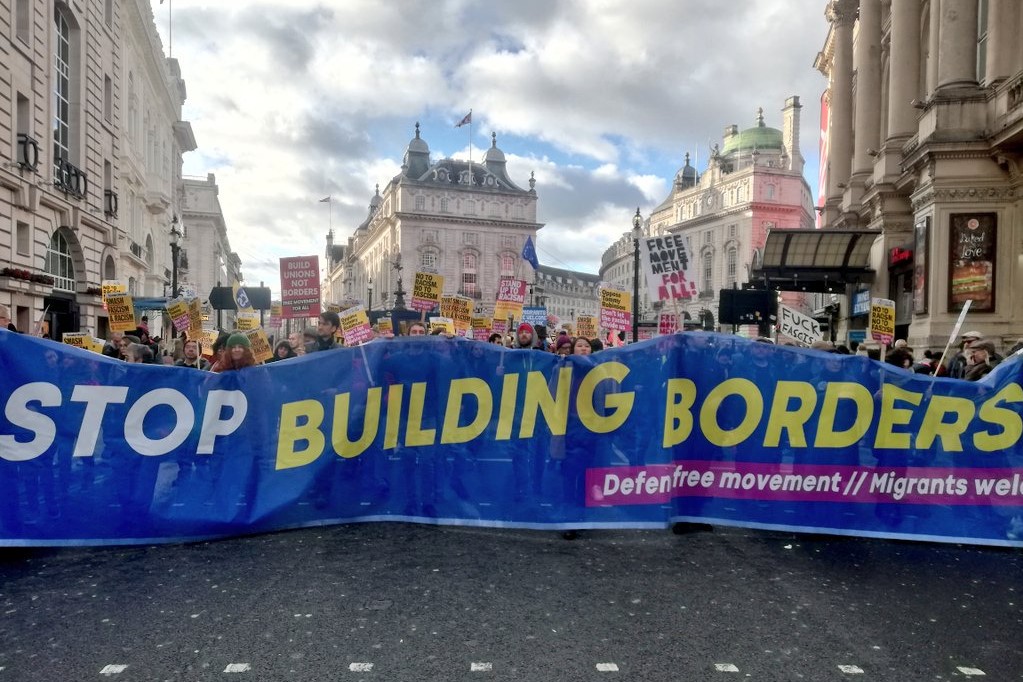 A crowd marches behind a banner reading 'STOP BUILDING BORDERS - Defend free movement - Migrants welcome'