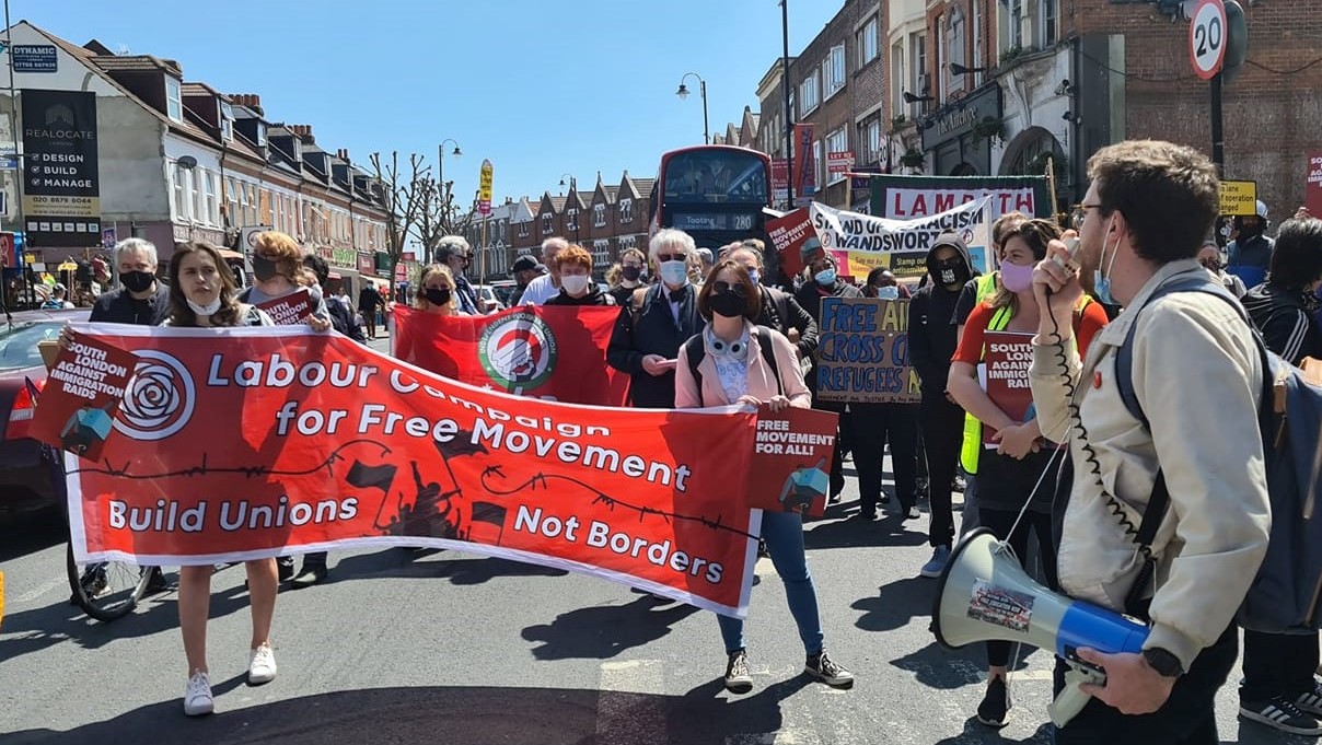 Protesters and trade union banners march down a street, towards the camera, behind the "Labour Campaign for Free Movement" banner. In the foreground, a person turns back towards the other protesters to chant through a megaphone.